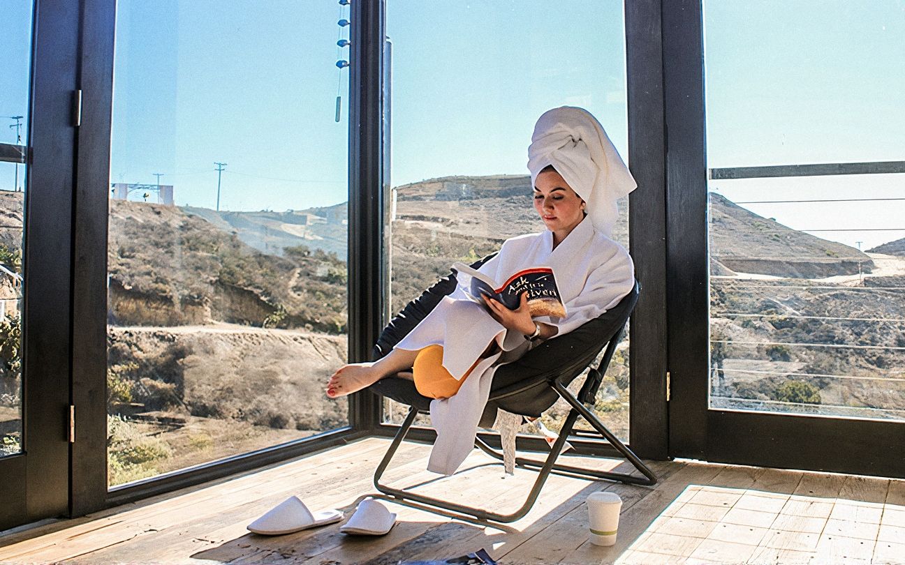 Huésped en bata leyendo en un sillón junto al ventanal de la Cabaña de Madera 01, con vista a las colinas