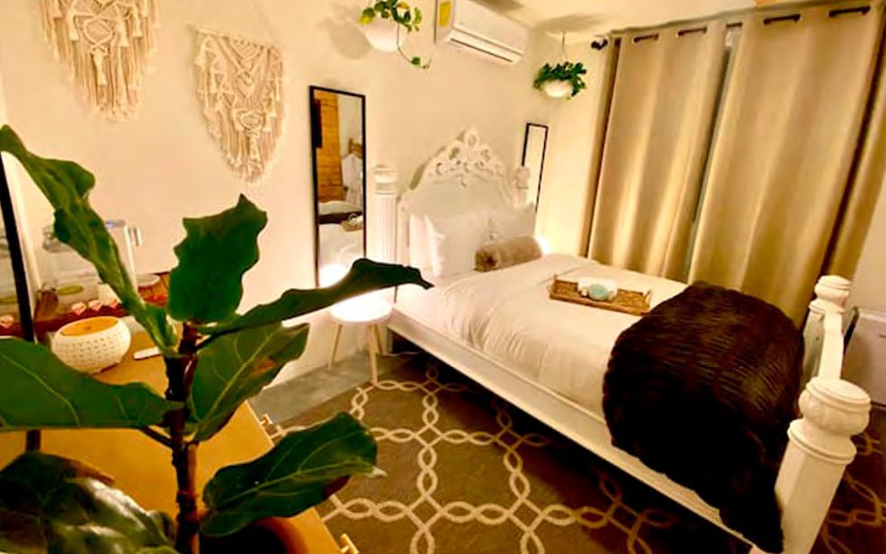 Habitación 03 de Casa Blanca con cama blanca de cabecera tallada, plantas, cortinas beige y tapete de cadenas